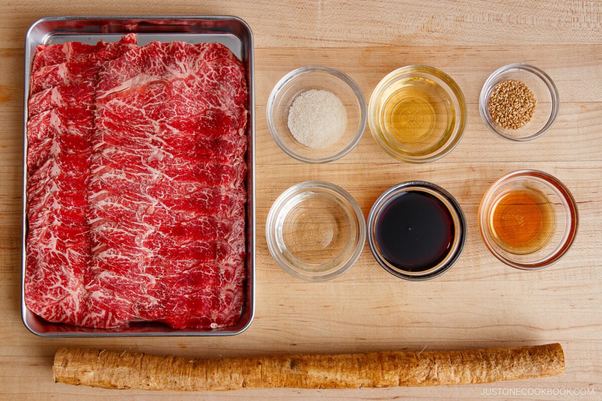 Thinly sliced marbled beef in a tray, six small bowls with various sauces and seasonings, and a long gobo root arranged neatly on a wooden surface&mdash;perfect ingredients for making Beef and Gobo Stir Fry.