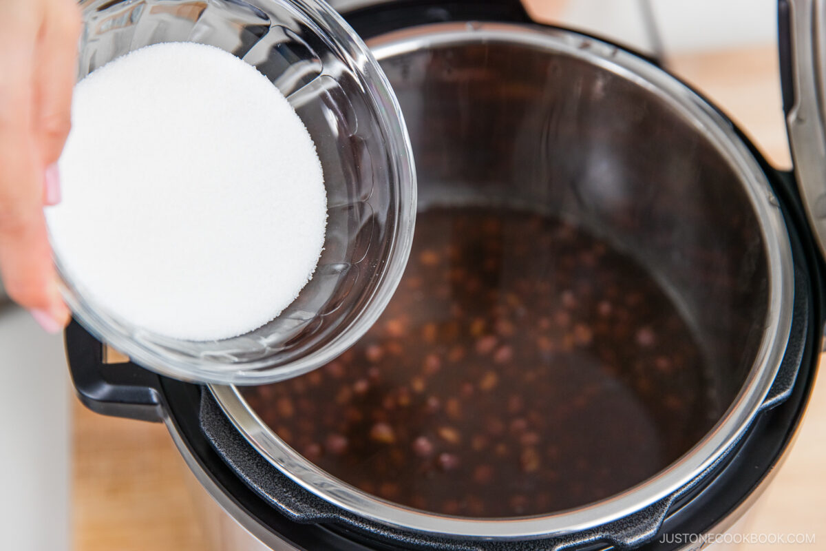 A hand pours a bowl of white sugar into an electric pressure cooker containing cooked red beans in water.
