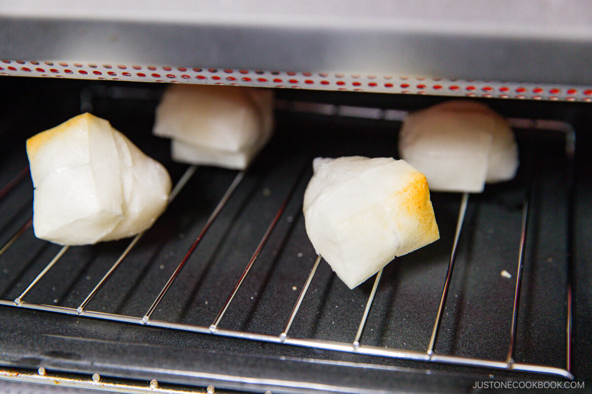 White mochi squares puff and toast on an oven rack, with slightly golden edges, inside an oven.