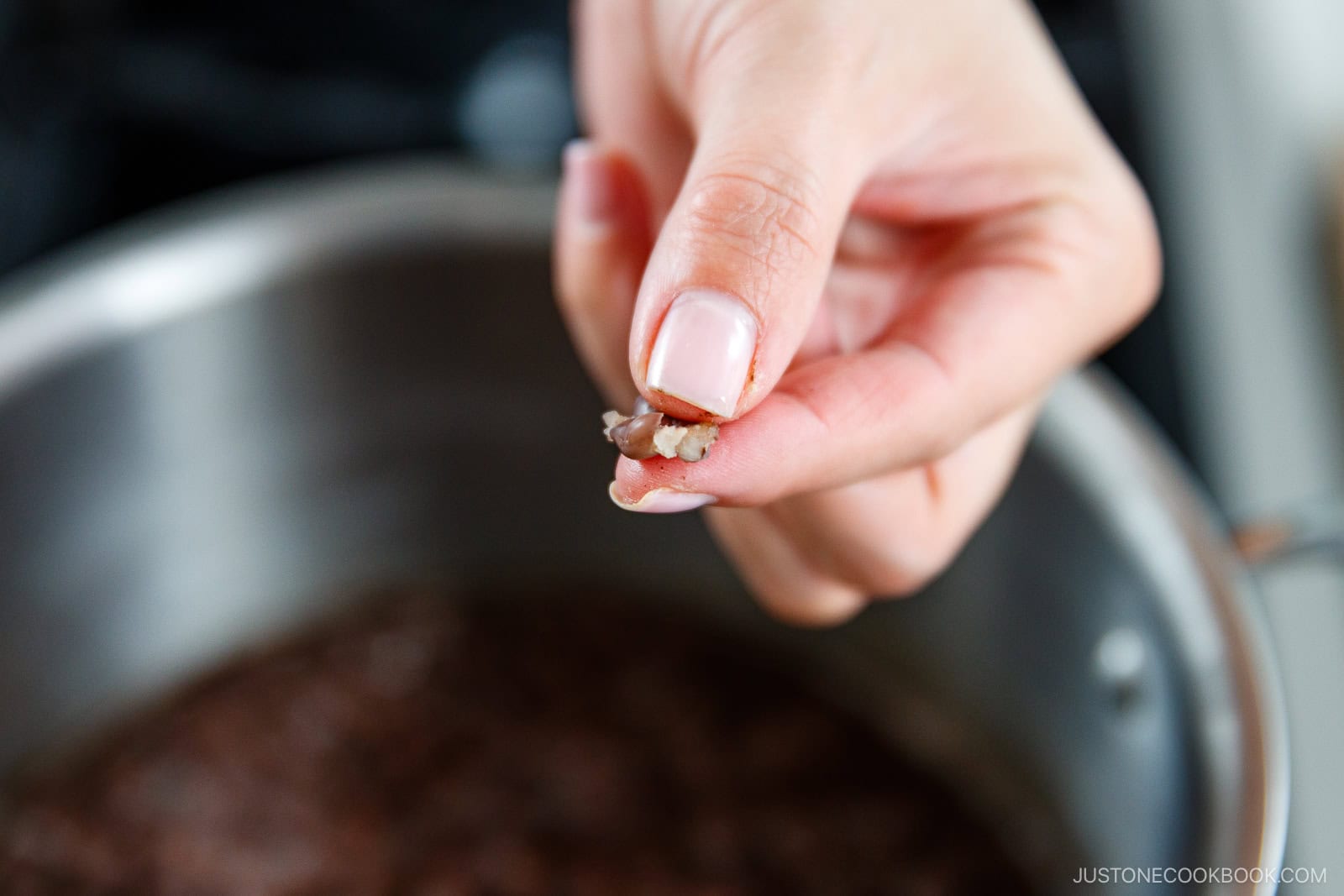 A close-up of a hand pinching a cooked red bean above a pot, checking its doneness. The background is slightly blurred, focusing attention on the hand and bean.