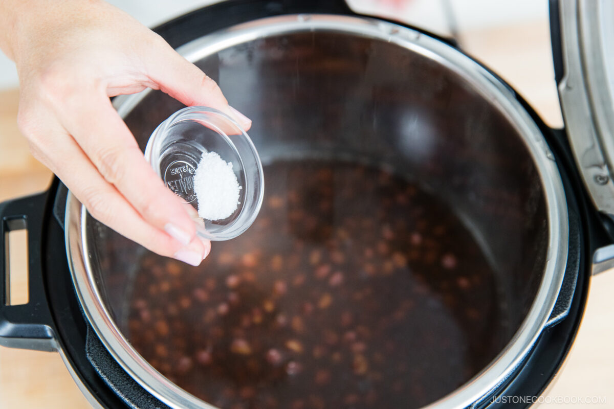 A hand is holding a small glass container of salt above an open electric pressure cooker filled with cooked beans and liquid.