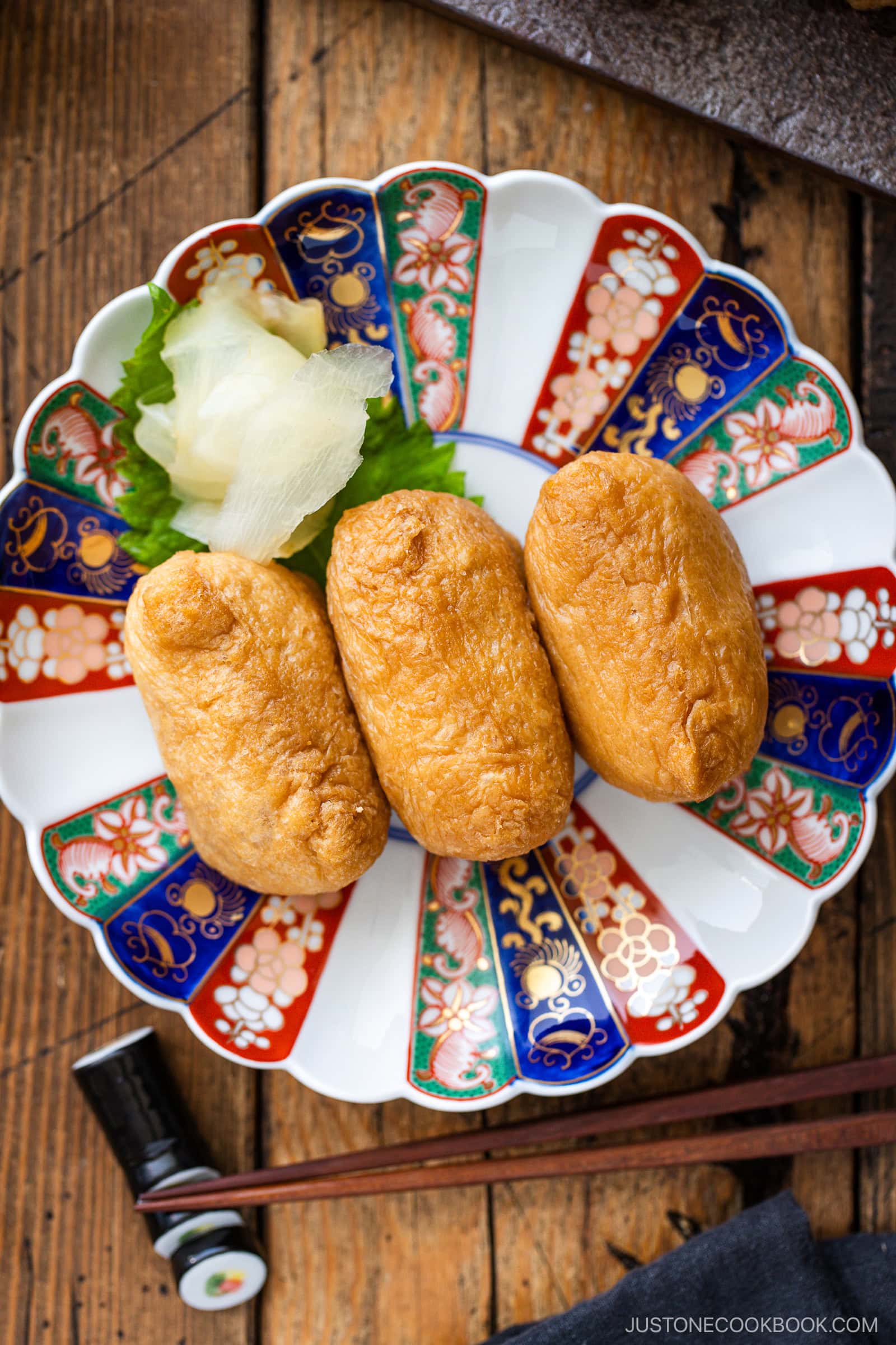 Three pieces of inari sushi are arranged on a decorative plate with pickled ginger and a shiso leaf, set on a wooden table with chopsticks and a small soy sauce dish nearby, ready to enjoy this classic inari sushi presentation.