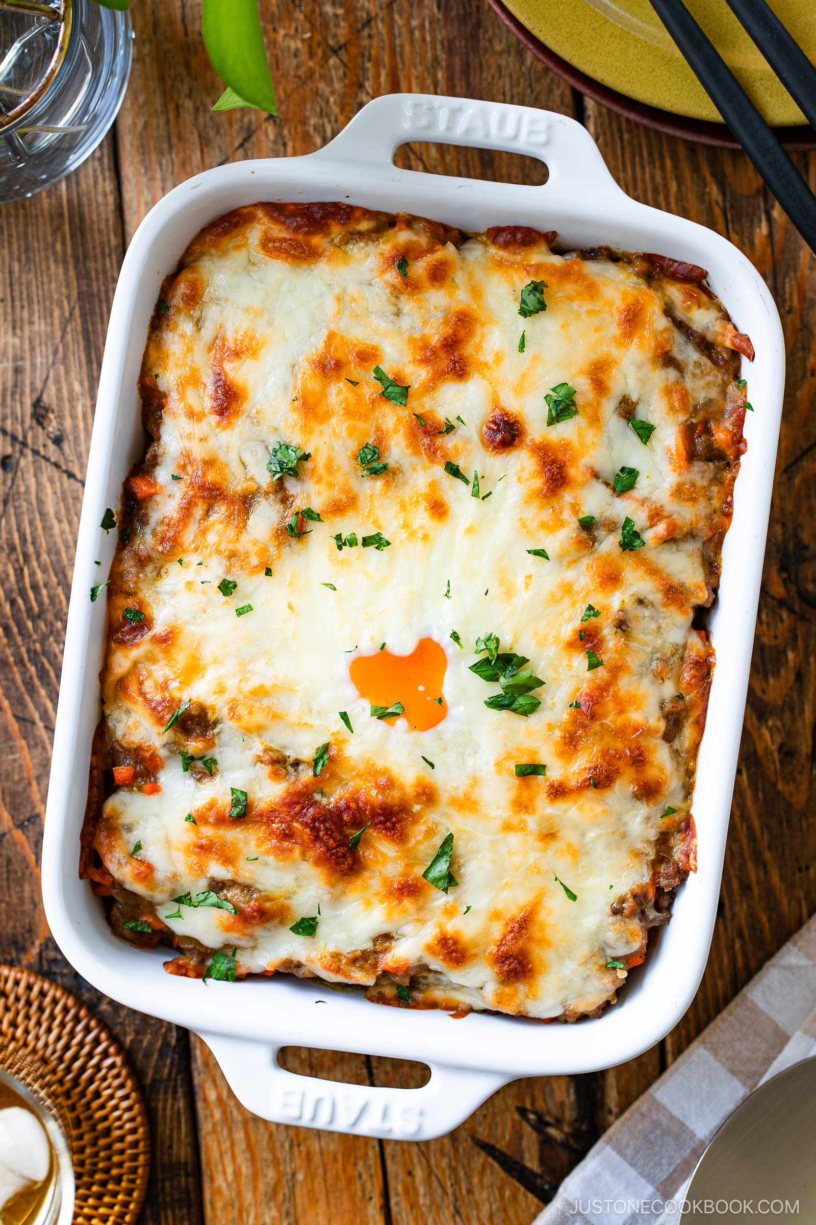 A white baking dish filled with a cheesy baked Yaki Keema Curry casserole, topped with melted golden-brown cheese and sprinkled with chopped herbs, sits on a wooden table set for a meal.