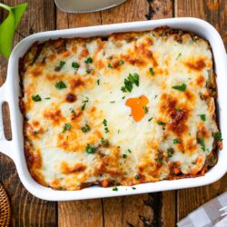 A baked Yaki Keema Curry casserole topped with melted cheese and parsley sits in a white dish on a wooden table, surrounded by plates, utensils, a napkin, and a glass of water.