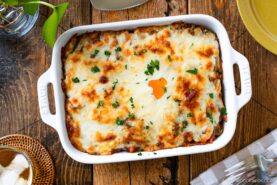 A baked Yaki Keema Curry casserole topped with melted cheese and parsley sits in a white dish on a wooden table, surrounded by plates, utensils, a napkin, and a glass of water.