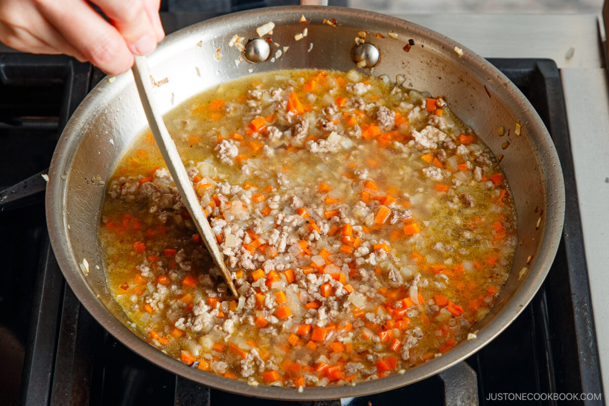 A hand stirs a savory mixture for Yaki Keema Curry&mdash;ground meat, diced carrots, and onions cooking in broth in a large stainless steel pan on a stovetop.