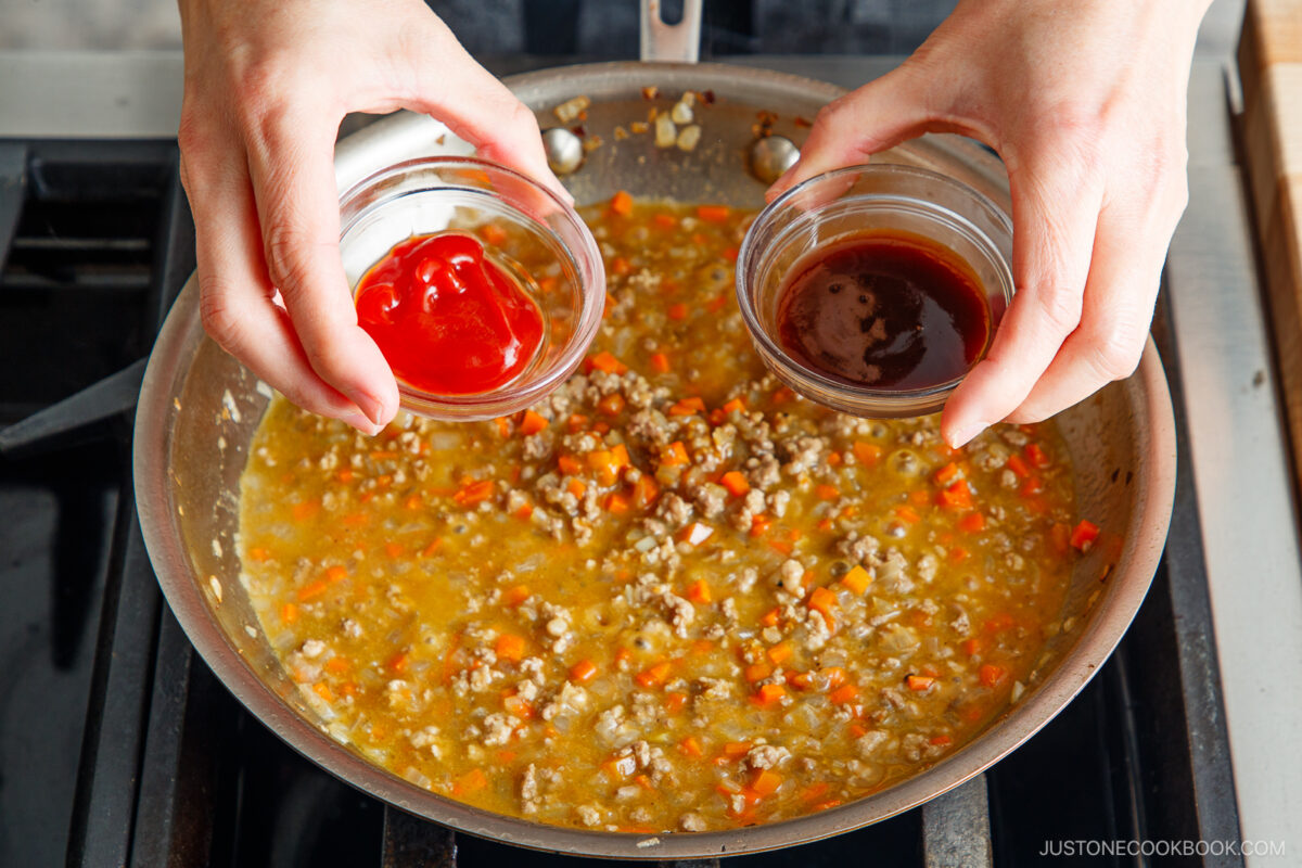 A person holds small bowls of ketchup and sauce above a pan of simmering ground meat with diced carrots and onions on a stovetop, preparing to add the sauces for Yaki Keema Curry.
