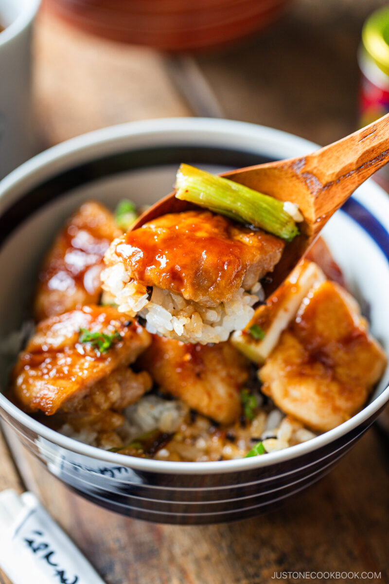 A wooden spoon holds a bite of yakitori don&mdash;glazed chicken, rice, and green onion&mdash;above a bowl filled with more saucy chicken and rice. The dish appears fresh and is garnished with chopped green onions.