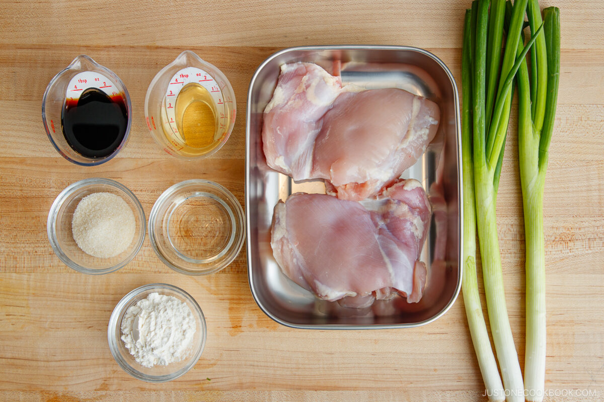 Raw chicken thighs on a metal tray, green onions, soy sauce, sake, sugar, flour, and cornstarch in glass bowls&mdash;all ingredients neatly arranged for a delicious yakitori don&mdash;set out on a wooden surface.