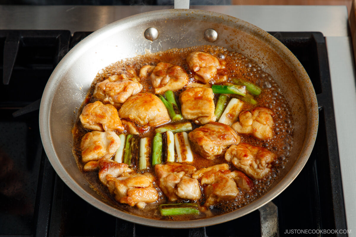 Chicken pieces and green onions are cooking in a skillet with a brown sauce on a stovetop. The golden chicken and bubbling sauce create a flavorful yakitori don-inspired dish.