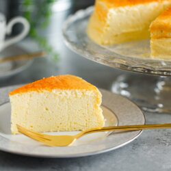 A slice of fluffy Japanese Cheesecake on a white plate with a gold fork, with the rest of the Japanese cheesecake recipe on a glass stand in the background. A cup of coffee and greenery are also visible.
