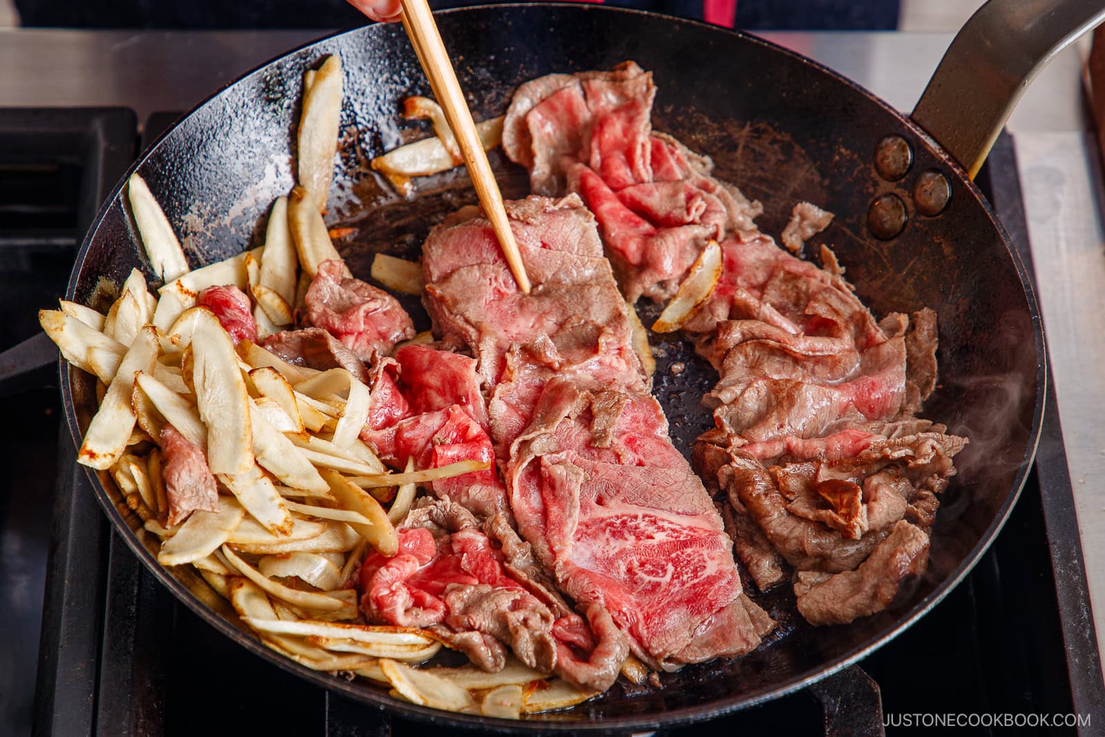 Thinly sliced beef and burdock root are being saut&eacute;ed for a savory Beef and Gobo Stir Fry in a black skillet. A hand uses chopsticks to cook the beef, which is partially browned and partially raw.
