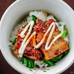 A bowl of white rice topped with glazed slices of meat, green vegetables, sesame seeds, and a drizzle of mayonnaise. Chopsticks are placed beside the bowl.