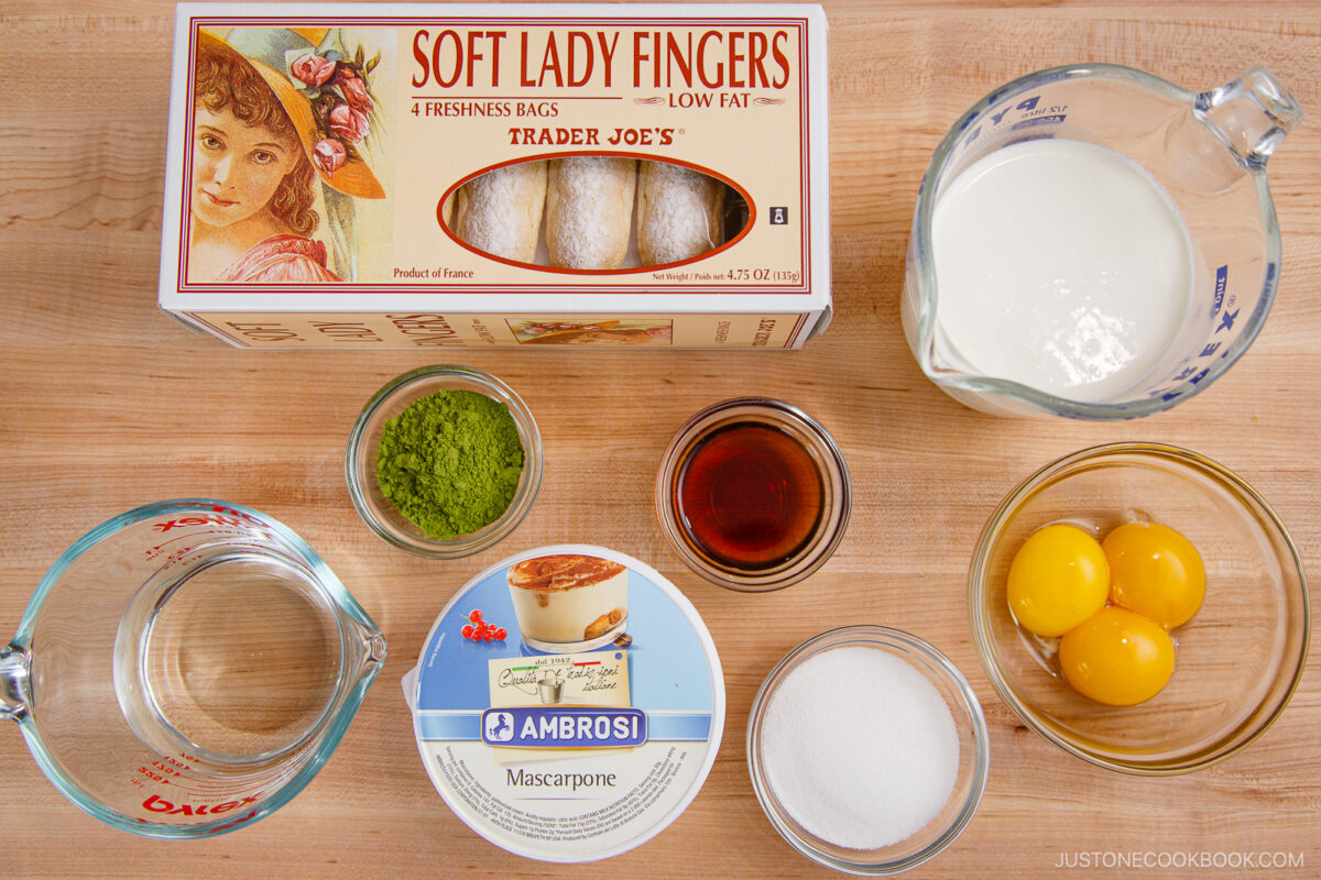 Top-down view of ingredients for matcha tiramisu on a wooden surface: ladyfingers, mascarpone, heavy cream, egg yolks, sugar, espresso, water, and matcha powder, each in separate bowls or containers.