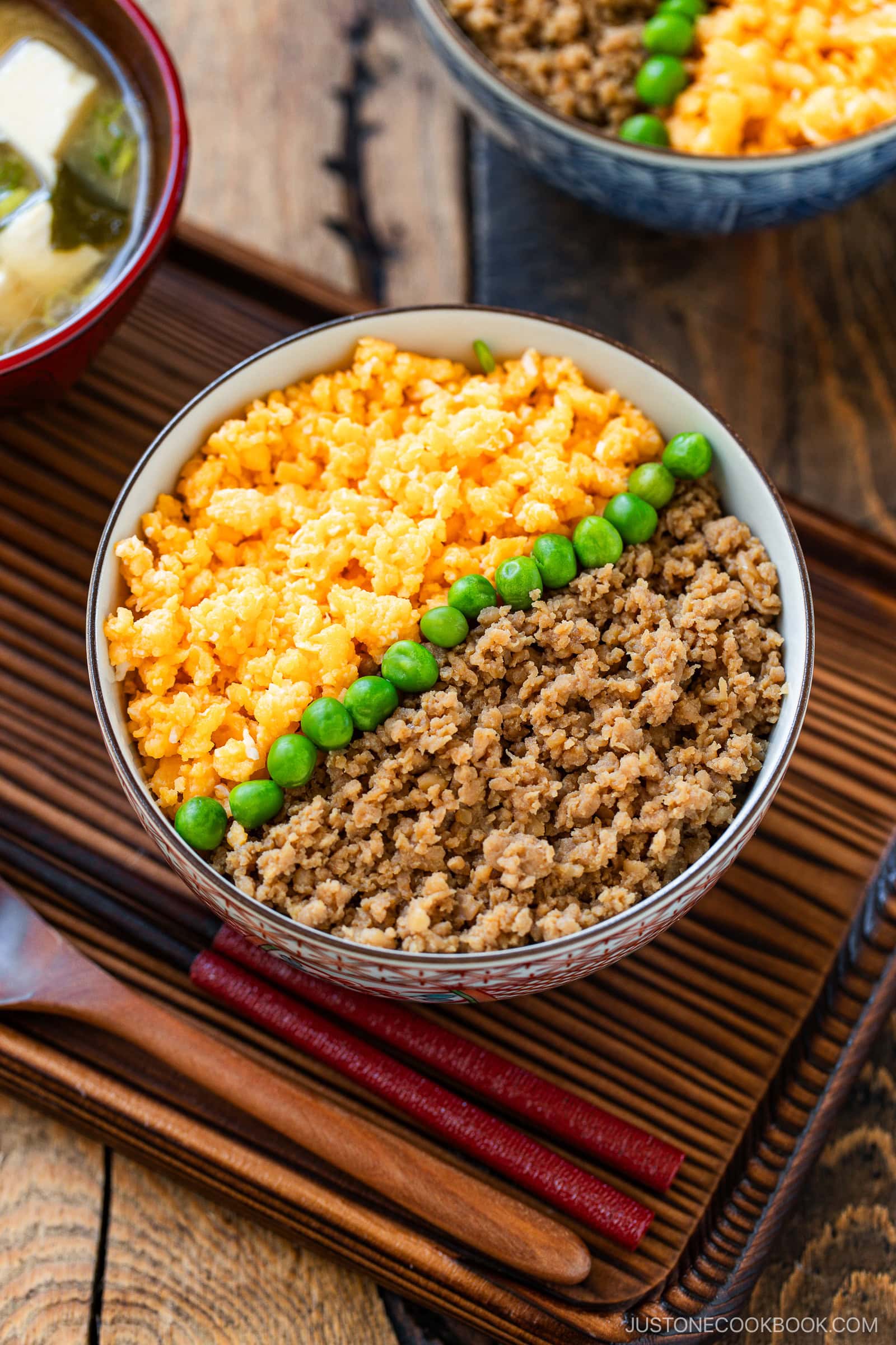 A bowl of soboro don (ground chicken bowl), featuring ground chicken, scrambled eggs, and green peas arranged in neat sections, sits on a tray next to a bowl of miso soup and a pair of wooden chopsticks.