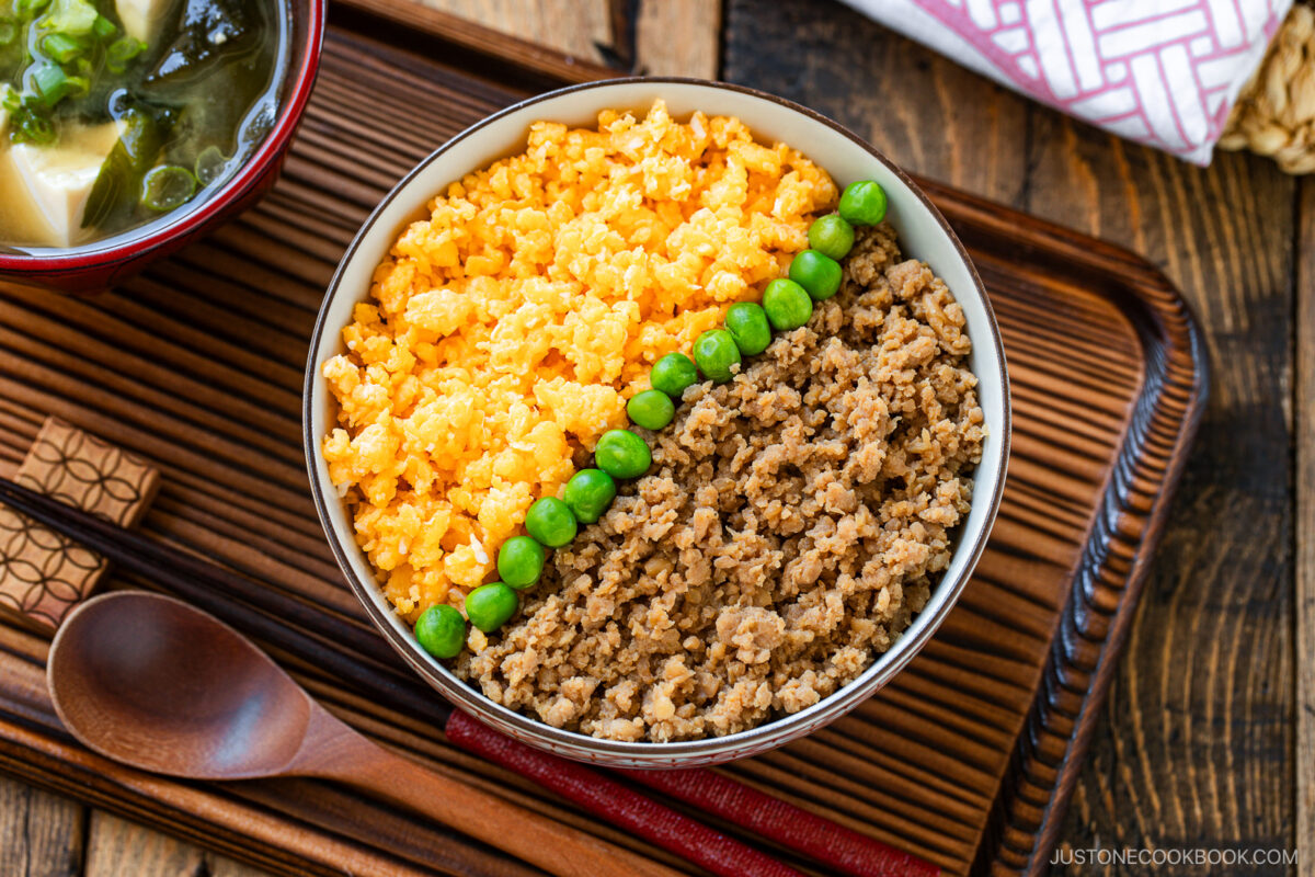 A bowl of soboro don (ground chicken bowl) with scrambled eggs and green peas arranged in neat sections, served on a wooden tray with a wooden spoon and a bowl of miso soup on the side.