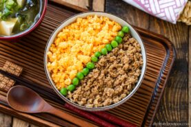 A bowl of soboro don (ground chicken bowl) with scrambled eggs and green peas arranged in neat sections, served on a wooden tray with a wooden spoon and a bowl of miso soup on the side.