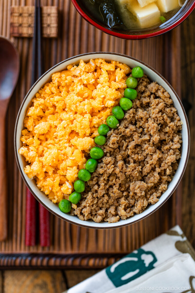A bowl of soboro don (ground chicken bowl) features ground meat on one side, scrambled egg on the other, and a row of green peas separating them, served on a wooden tray with chopsticks and miso soup.
