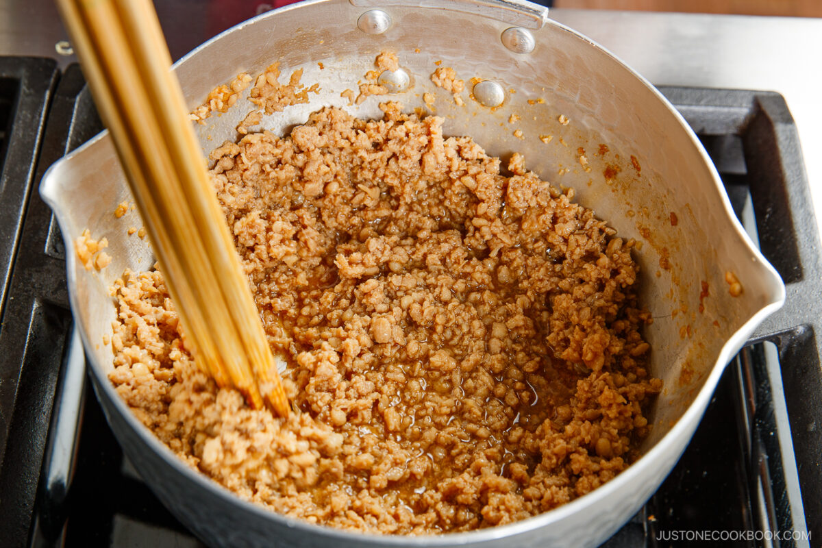 Ground meat, similar to what's used in soboro don (ground chicken bowl), is being cooked in a pot with brown sauce as uncooked spaghetti noodles are added. The savory mixture simmers on the stovetop, ready for a quick and flavorful meal.