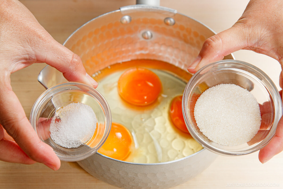 A close-up of hands holding two small bowls, one with sugar and one with salt, over a pot containing cracked eggs and a liquid mixture for making soboro don (ground chicken bowl).