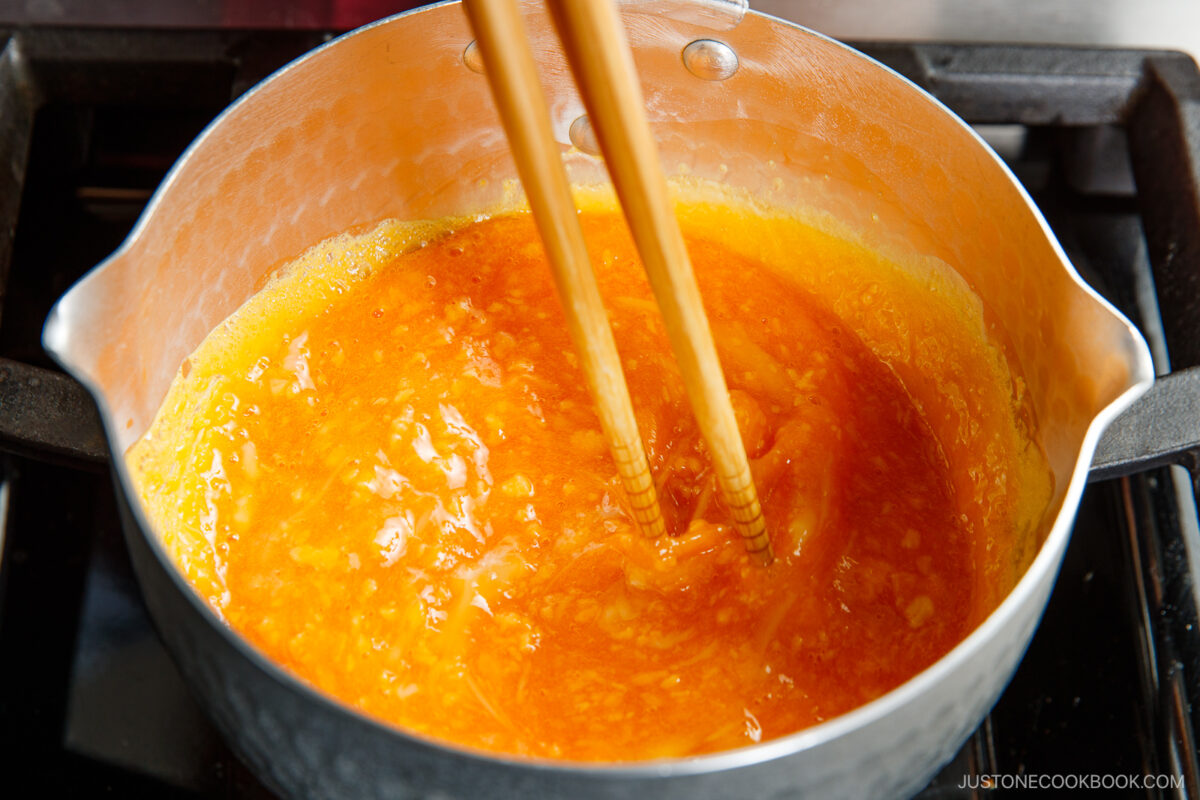 A close-up of a metal pot on a stovetop containing bright yellow-orange scrambled eggs for soboro don, being stirred with a pair of wooden chopsticks.