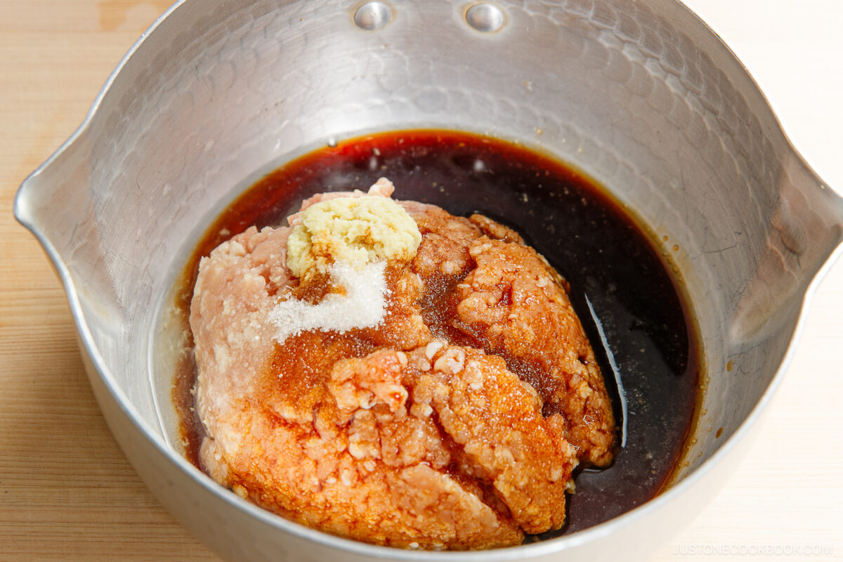 Raw ground meat in a metal bowl with soy sauce, grated ginger, and salt sprinkled on top, ready to be mixed for cooking soboro don (ground chicken bowl).