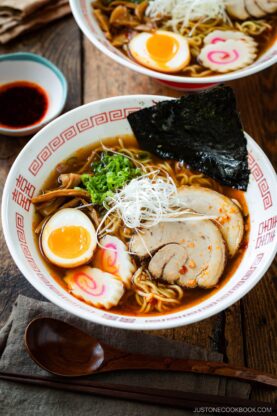 A bowl of spicy shoyu ramen with sliced pork, soft-boiled egg, narutomaki, nori seaweed, green onions, mushrooms, and noodles in broth, served on a wooden table with a wooden spoon and a small dish of sauce.