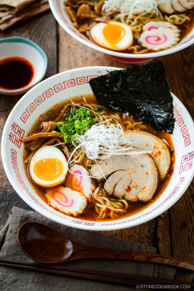 A bowl of spicy shoyu ramen with sliced pork, soft-boiled egg, narutomaki, nori seaweed, green onions, mushrooms, and noodles in broth, served on a wooden table with a wooden spoon and a small dish of sauce.