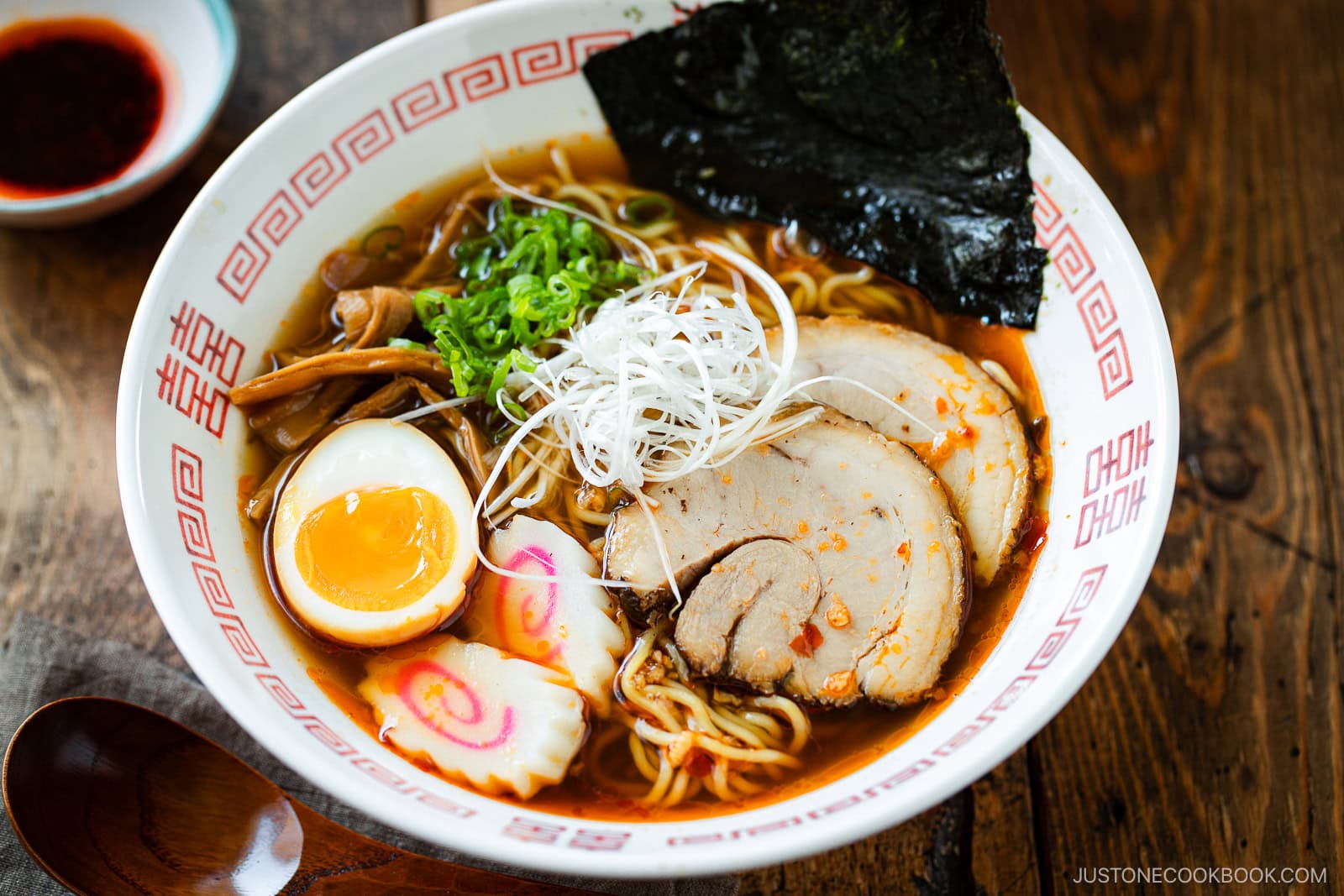 A bowl of spicy shoyu ramen topped with sliced pork, half a soft-boiled egg, green onions, seaweed, bamboo shoots, fish cake slices, and white shredded onions, served in a patterned bowl on a wooden table.