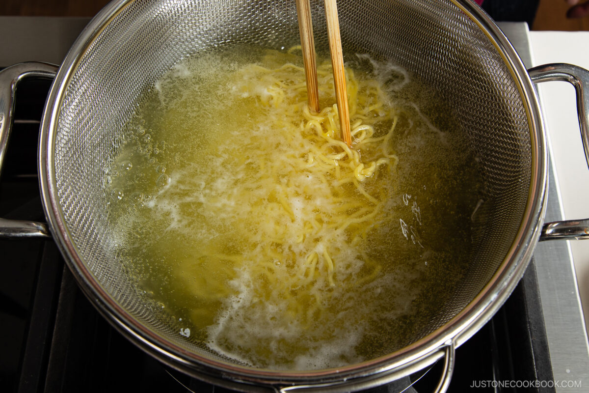 Spicy shoyu ramen noodles cooking in boiling water in a metal pot, stirred with wooden chopsticks on a stovetop.