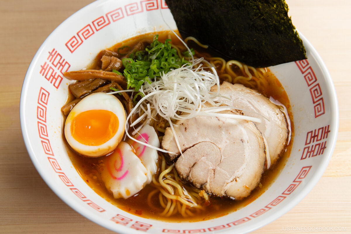 A bowl of spicy shoyu ramen with sliced pork, half a soft-boiled egg, narutomaki, bamboo shoots, green onions, shredded white onions, and a sheet of nori in a patterned bowl on a wooden table.