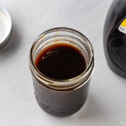 An open glass jar filled with dark yakisoba sauce, made from a classic yakisoba sauce recipe, sits on a white surface next to its lid and a bottle labeled “Yakisoba Sauce.”.
