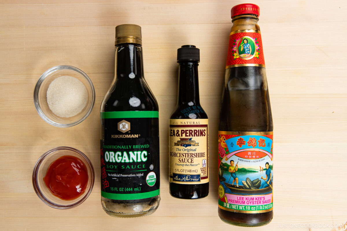 Three bottles—soy sauce, Worcestershire sauce, and oyster sauce—are lined up on a wooden surface with small bowls of granulated sugar and ketchup to the left, all classic ingredients for a homemade yakisoba sauce recipe.