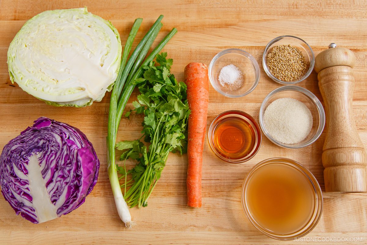 Green and purple cabbage, green onions, cilantro, a carrot, salt, sesame seeds, pepper, sugar, rice vinegar, and soy sauce are arranged neatly on a wooden cutting board—perfect ingredients for preparing a fresh Asian Coleslaw with Sesame Dressing.