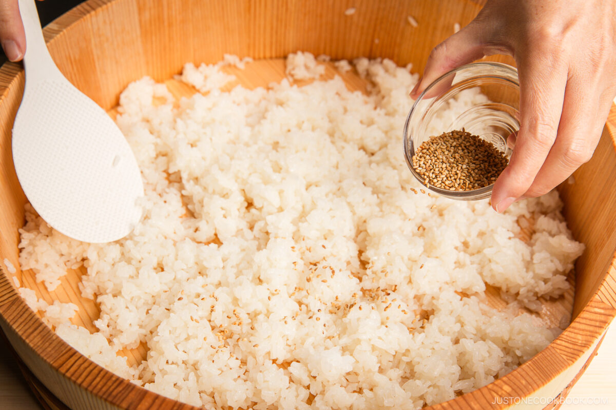 A hand sprinkles sesame seeds from a small glass bowl onto cooked white rice in a wooden bowl, perfect for making inari sushi. A white rice paddle rests on the side of the bowl.
