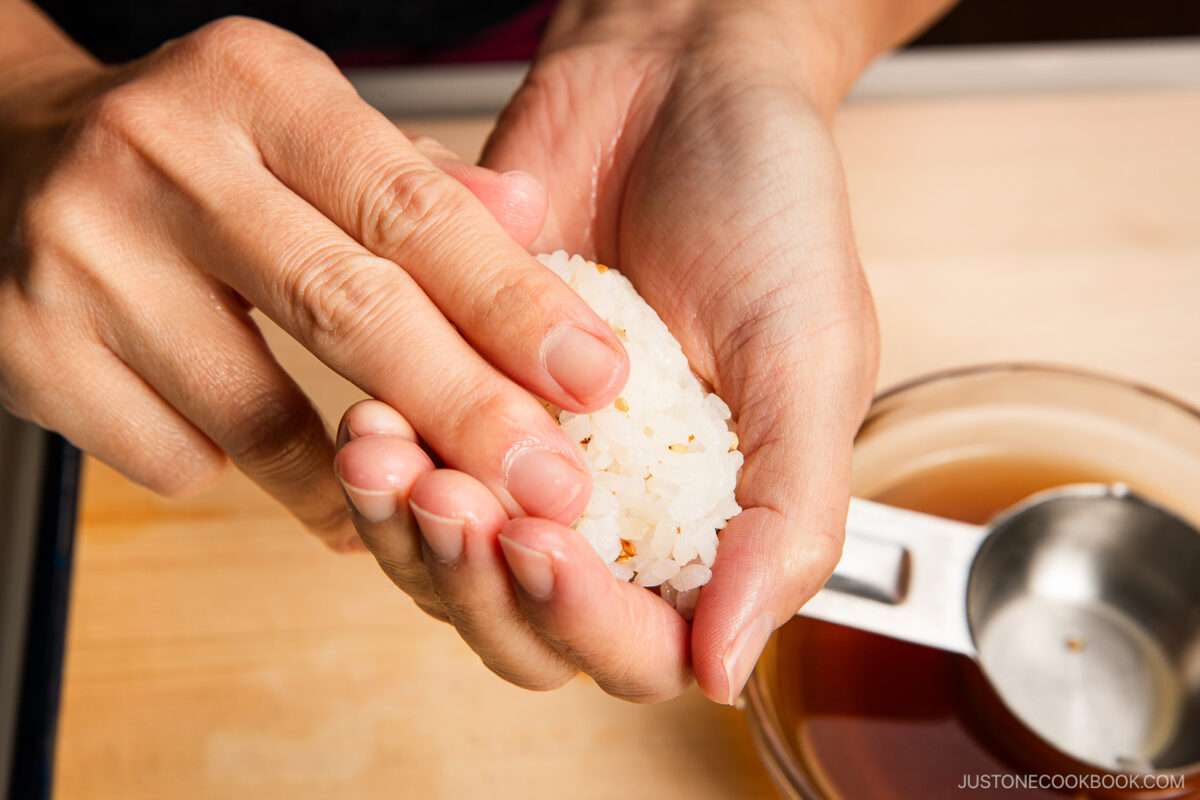 Close-up of hands shaping white rice into a small oval for inari sushi, with a bowl of liquid and a metal measuring cup nearby on a wooden surface.