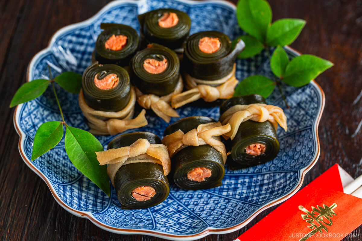 A blue patterned plate holds neatly arranged Kobumaki (Salmon Kombu Roll)—kelp seaweed rolls with salmon filling, tied with a light-colored gourd strip and garnished with green leaves. Red envelope chopsticks and a napkin are beside the plate.