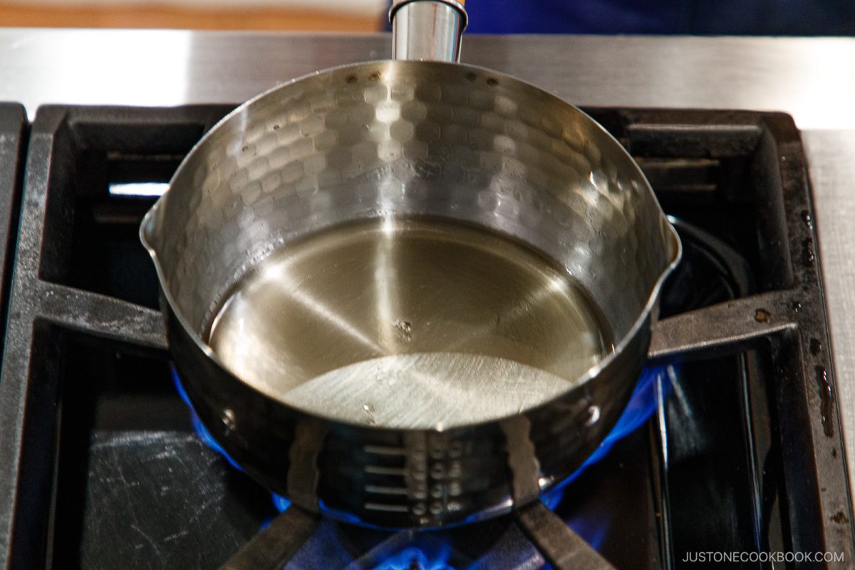 A stainless steel pot with oil and a splash of nikiri mirin heats on a gas stove, with blue flames visible beneath the pot.