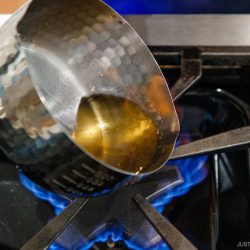 A metal pot with oil and nikiri mirin is tilted over a blue gas flame on a stove burner.