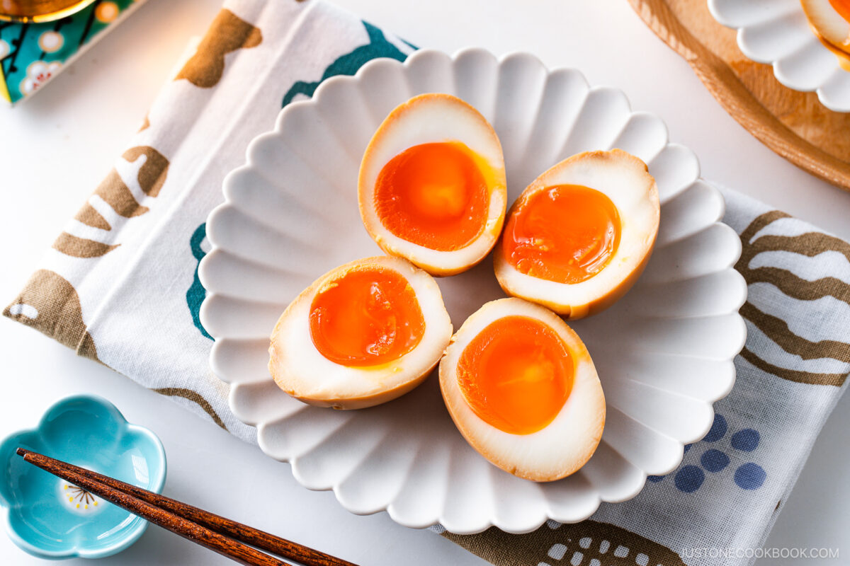 Four ramen eggs, halved to reveal their soft, runny yolks, are arranged on a white scalloped plate set on a patterned cloth. A pair of wooden chopsticks and a blue dish complete this inviting ramen eggs presentation.