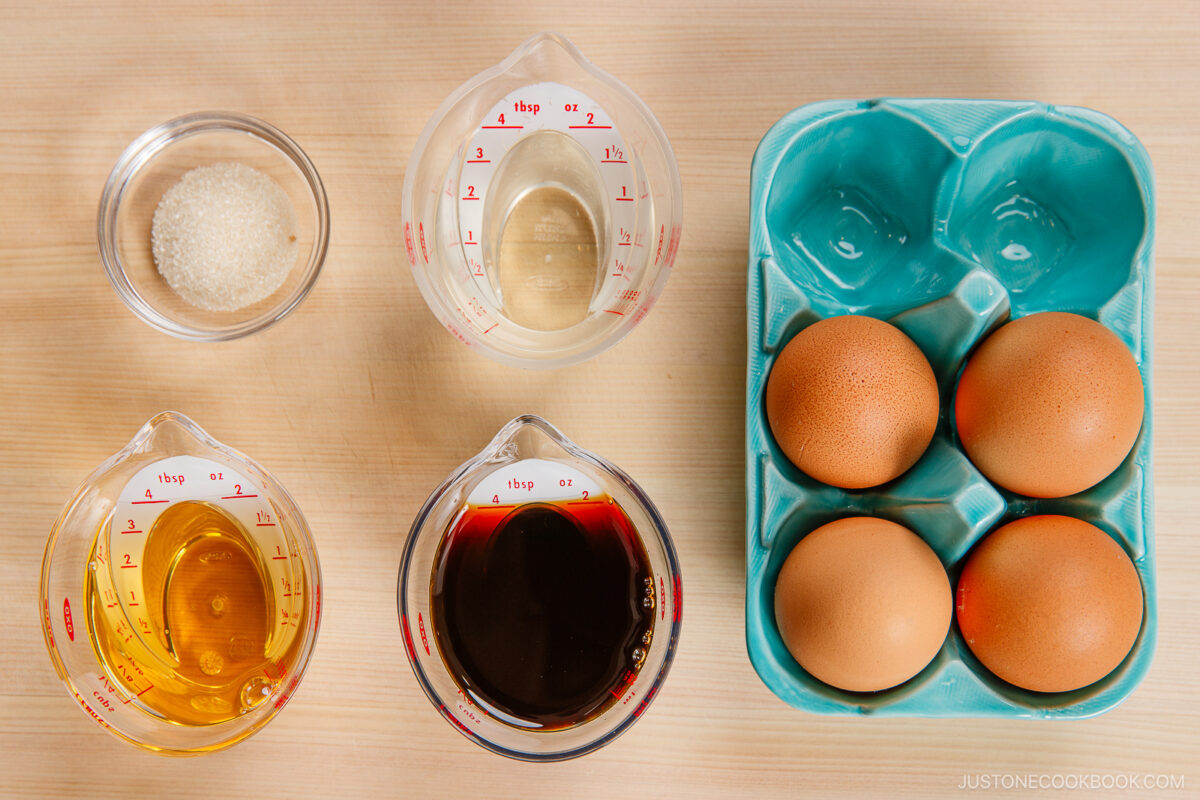 Top-down view of four brown eggs in a blue carton, perfect for making ramen eggs, and four clear containers holding sugar, water, oil, soy sauce, and broth&mdash;all neatly arranged on a wooden surface.