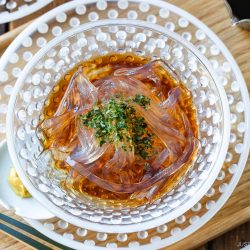 A glass bowl of clear Japanese kanten noodles in brown sauce, garnished with green herbs, sits on a dotted plate with chopsticks and a spoon on a wooden tray.