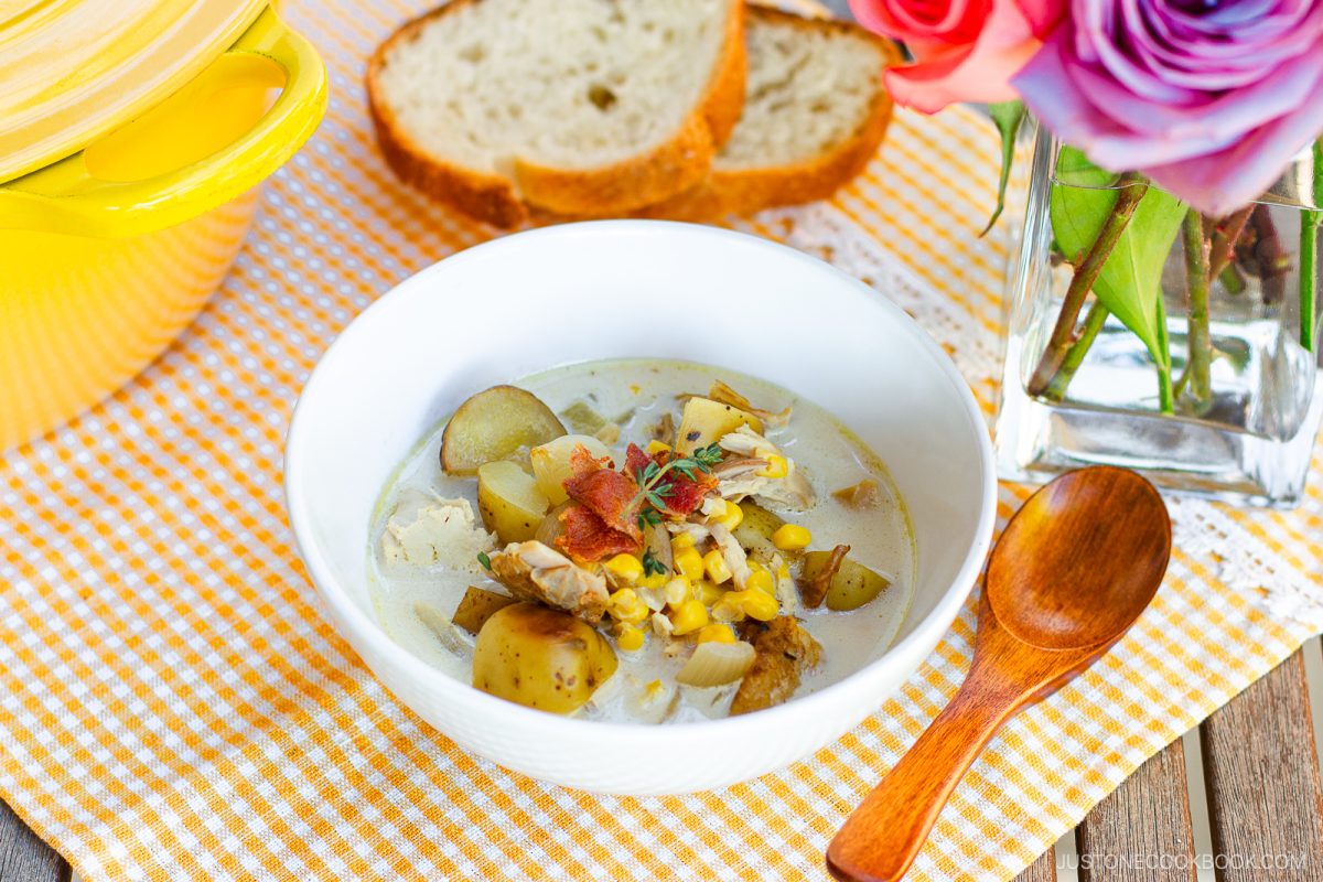 A bowl of creamy Chicken Corn Chowder with tender chicken and potato, topped with corn and bacon, sits on a yellow gingham cloth next to a wooden spoon, bread slices, a yellow pot, and a vase of colorful flowers.