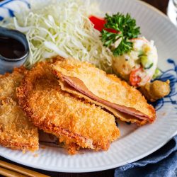 A plate with breaded, fried pork cutlets—reminiscent of Ham Katsu from Midnight Diner—shredded cabbage, potato salad, a dollop of mustard, and a small dish of dark sauce. Chopsticks and a drink are beside the plate.