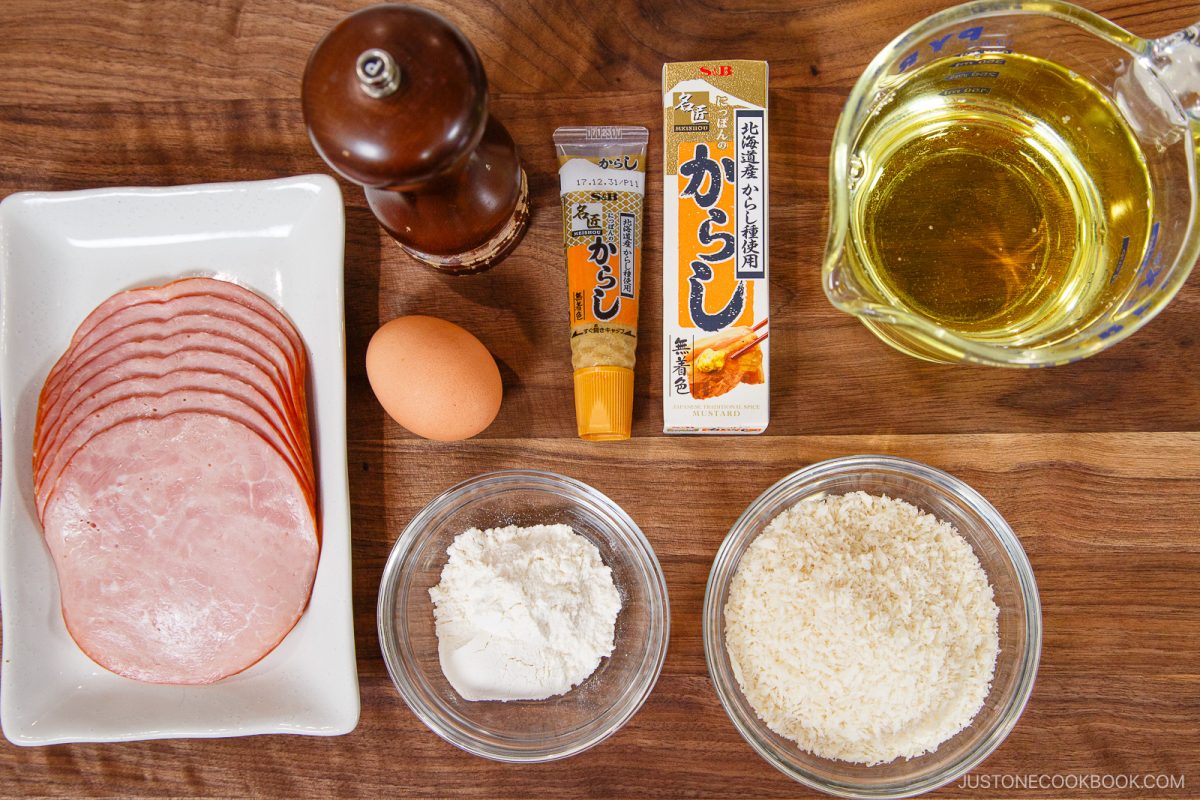 Sliced ham, an egg, flour, panko breadcrumbs, a pepper grinder, karashi mustard, and a measuring cup of oil are arranged neatly on a wooden surface—perfect for recreating Ham Katsu (Midnight Diner) at home.