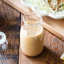 A glass jar filled with creamy sesame dressing sits on a wooden table, with its lid nearby and a plate of shredded cabbage salad blurred in the background.