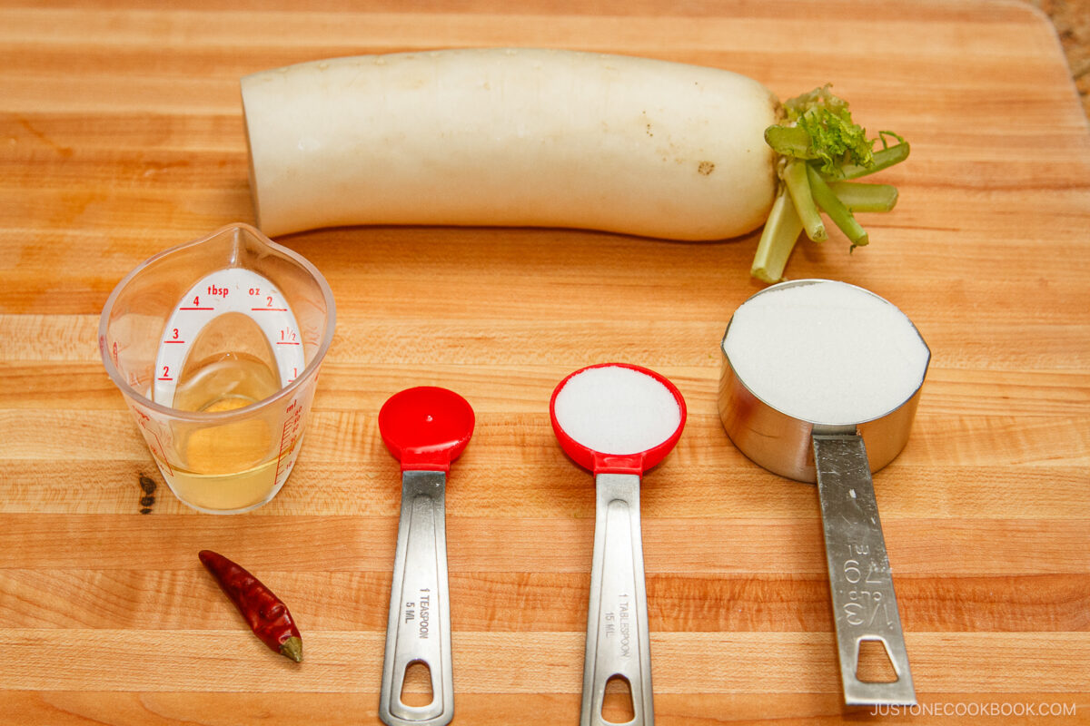 A daikon radish, a small cup of liquid, a dried red chili, a tablespoon of salt, a tablespoon of sugar, and a cup of white sugar are arranged on a wooden cutting board—perfect ingredients for making pickled daikon.