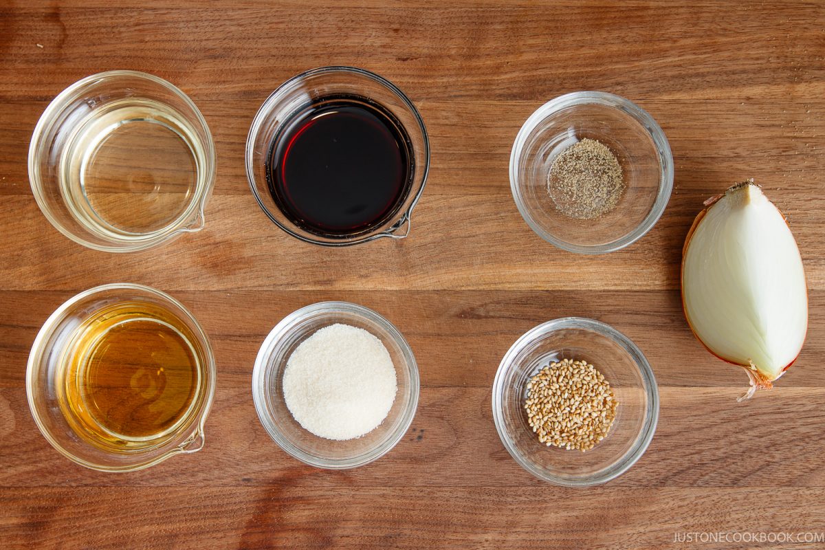 Six small bowls with oil, soy sauce, pepper, sesame oil, sugar, and sesame seeds are arranged on a wooden surface, alongside half a yellow onion.