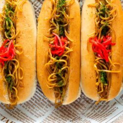 Five Yakisoba Pan just like on Midnight Diner—Japanese hot dog buns filled with stir-fried noodles, green herbs, and red pickled ginger—are arranged side by side on a newspaper-lined surface.