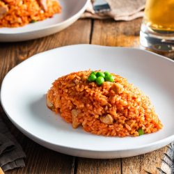 A white plate holds a serving of Japanese Chicken Rice (Midnight Diner), topped with green peas. A fork, napkin, beer can, and glass are nearby on a wooden table.